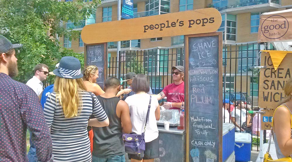 Group of people standing in line for ice pop vendor at Brooklyn outdoor market.